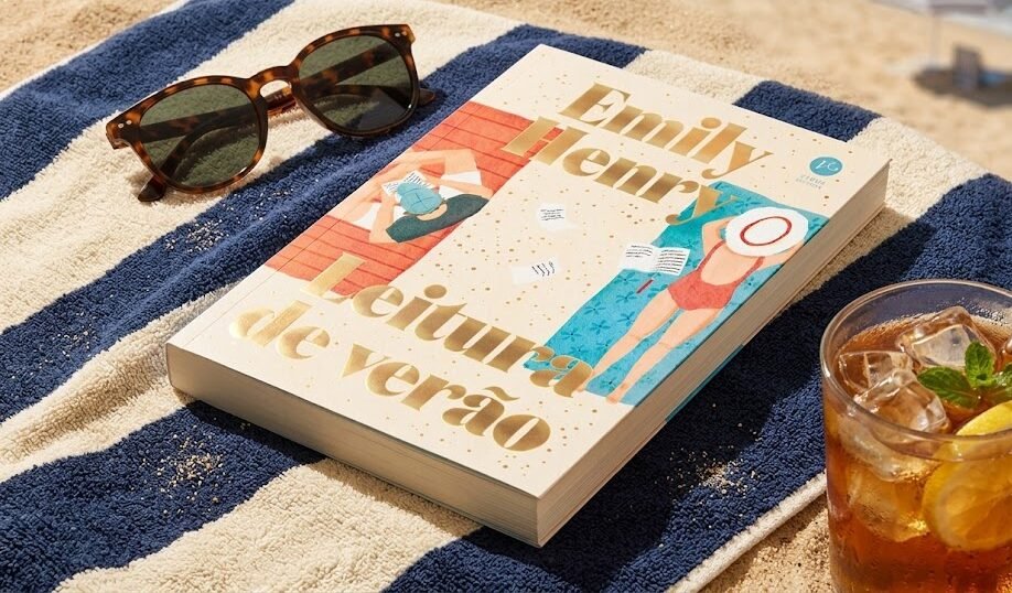 Livro Leitura de Verão de Emily Henry apoiado em uma toalha listrada na areia da praia, ao lado de óculos de sol chiques e um chá gelado refrescante.